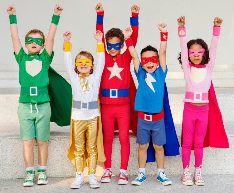 Five children in superhero costumes with arms raised, smiling outdoors.