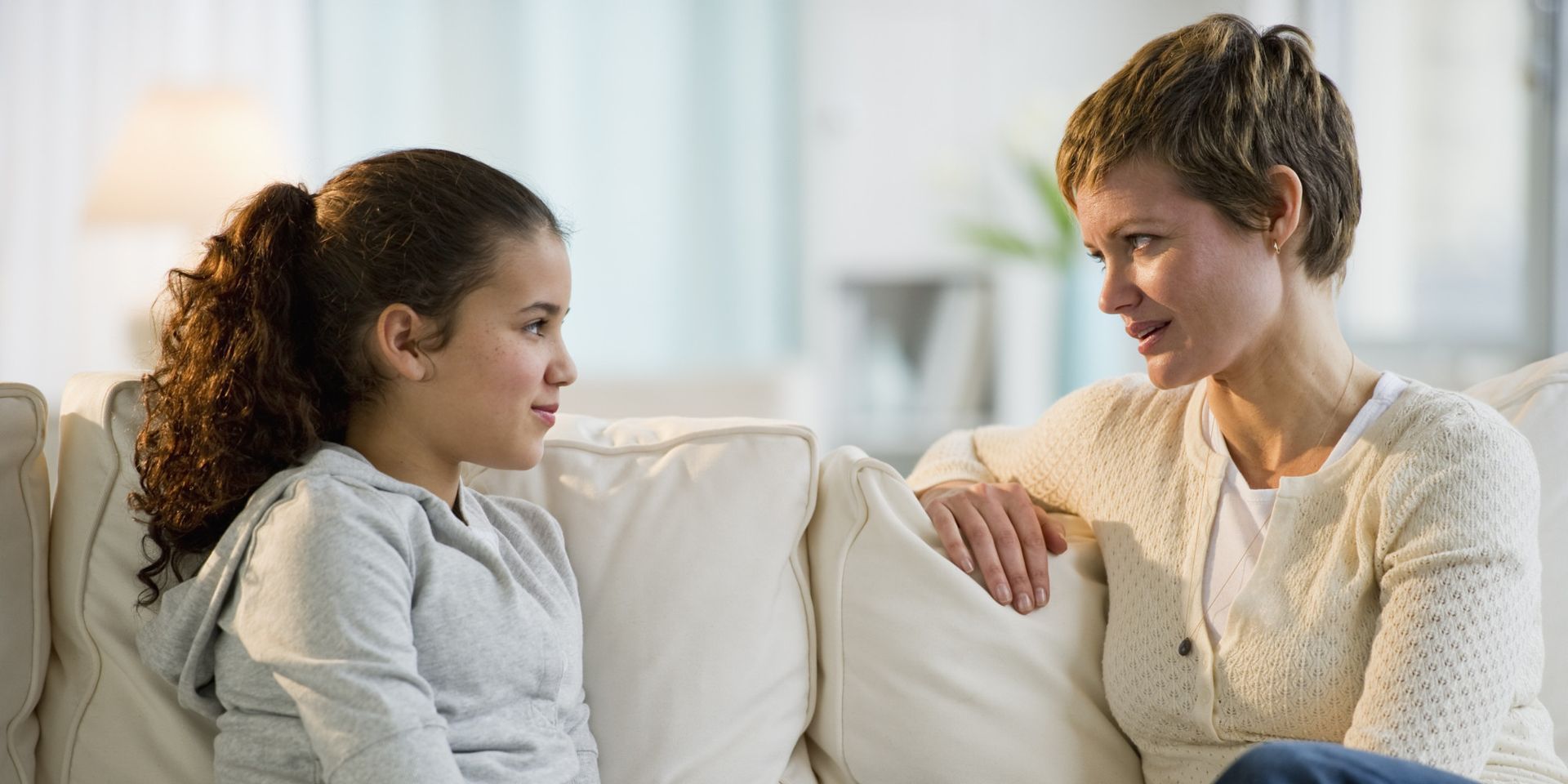 A woman and a girl sitting on a couch, talking. The woman has short hair and the girl has long curly hair.