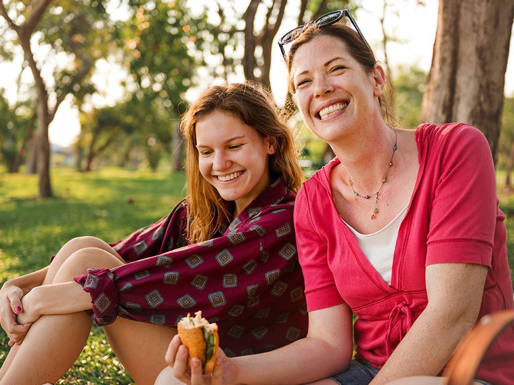 Woman and girl smiling, eating outdoors. Lush green background with trees, both in casual clothes.