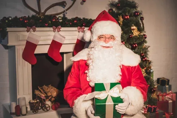 Santa Claus sitting by a fireplace with a gift, Christmas tree, and stockings; festive holiday scene.