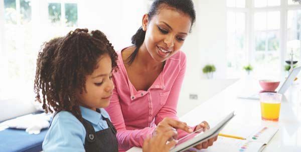 Mother and child using a tablet together in a bright kitchen. The child points, the mother smiles.