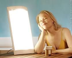 Woman using a light therapy lamp. Yellow top, blonde hair, resting on hand, smiling. Blue background, wooden table.