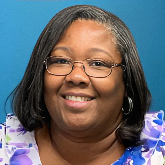 Woman with glasses smiling, wearing a floral blouse, against a blue background.