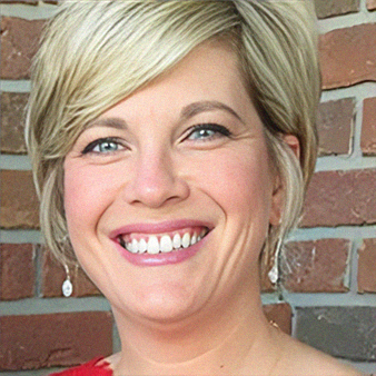 Blonde woman with blue eyes smiles at the camera, wearing earrings, in front of a brick wall.