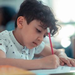 Boy with dark hair writing with a red pencil at a desk, focused, white shirt.