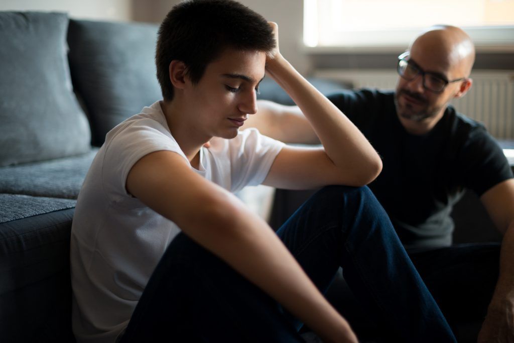Teenager sits, head in hand, looking sad, while a man comforts him.