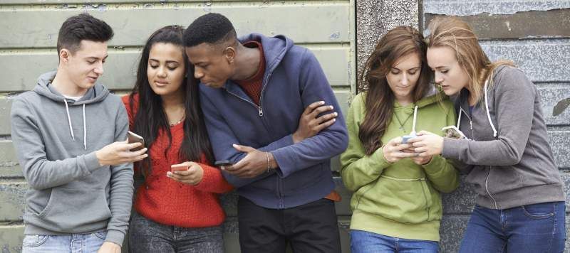 Group of five teens looking at their phones together, leaning against a weathered wall.