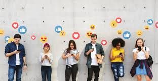 People looking at phones, standing in front of a gray wall with floating social media icons: likes, hearts, and emojis.