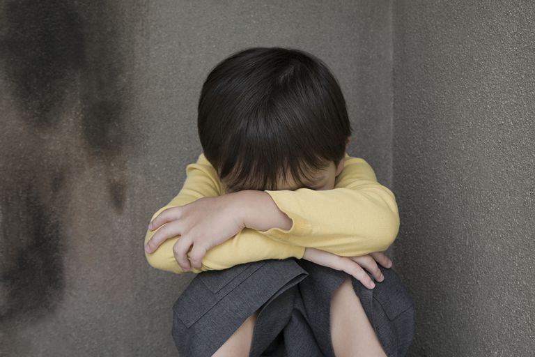 Child with head down, hugging knees, sitting in a corner, looking sad, wearing yellow shirt and grey pants.