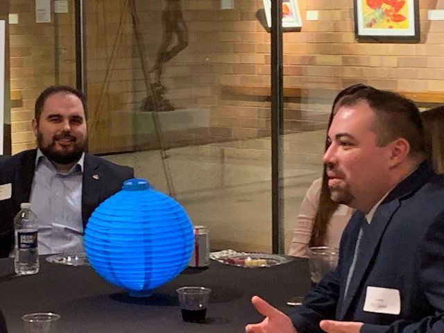 Two men in suits at a table with a blue lantern. One speaks, the other smiles. Brick wall and art visible.