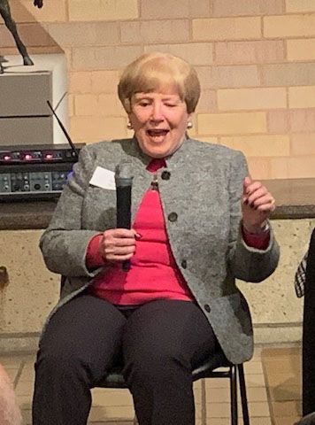 Woman with short blonde hair speaks into a microphone, wearing a gray jacket, red shirt, and black pants; sitting.