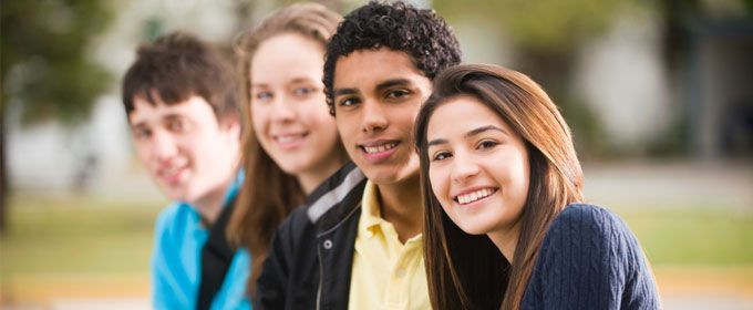 Four smiling teenagers in a row, outdoors, with varying skin tones and hair colors.