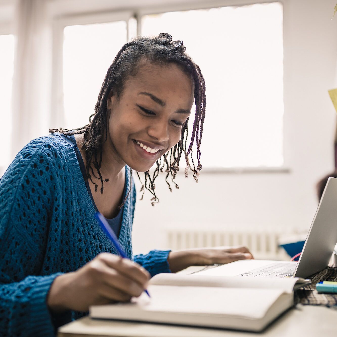 Smiling woman with locs writes in a notebook, using a laptop in a well-lit room.