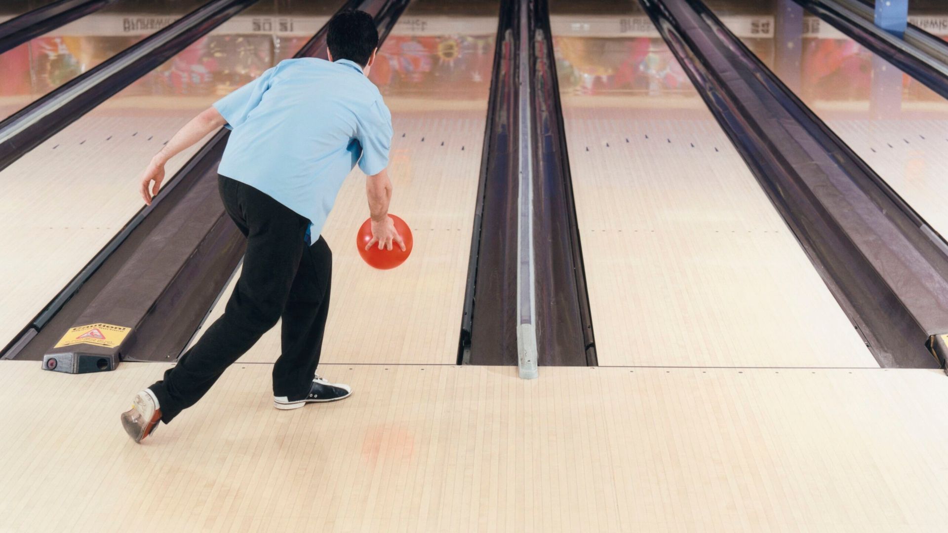 Bowler in a blue shirt, bowling down the lane, holding a red ball.