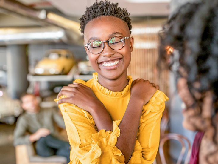 Woman with glasses, yellow shirt, hugging herself and smiling. Cafe setting, cars in the background.
