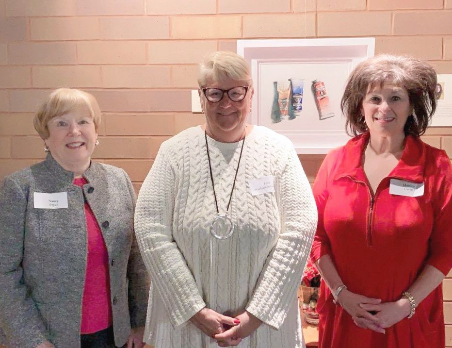 Three women stand together. One wears a gray jacket, pink shirt, and has a name tag. Another wears a cream sweater. The third wears red and has a name tag.