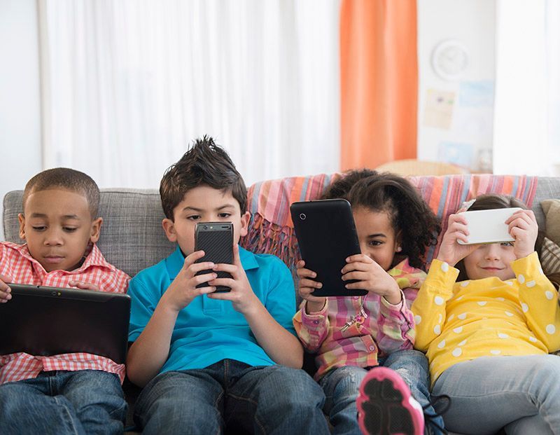 Four children of diverse ethnicities absorbed in using tablets and phones on a couch.