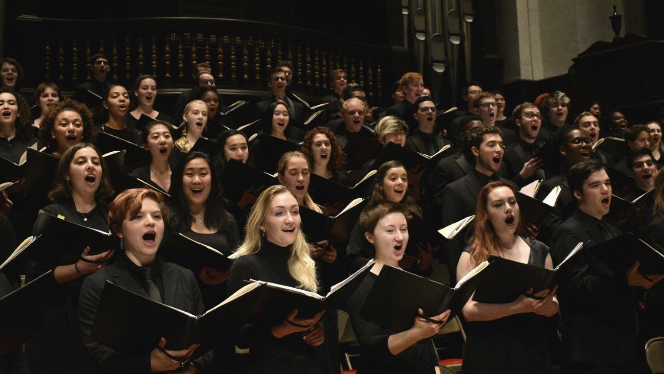 Choir singing with open mouths and sheet music, wearing black in a dark hall.