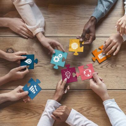 Hands holding puzzle pieces with icons on a wooden table, symbolizing collaboration and problem-solving.