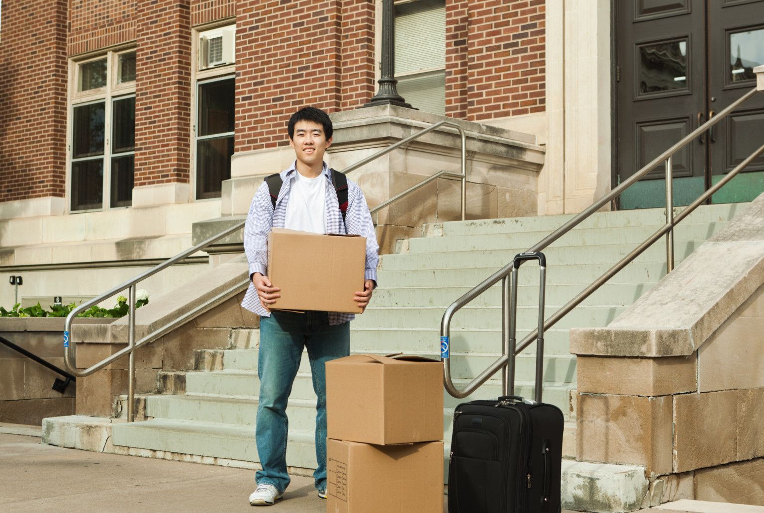 Young Asian man moves boxes and luggage outside a brick building.