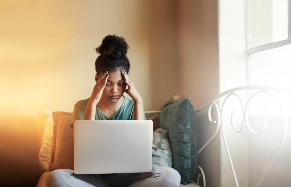 Woman with curly hair, hands on temples, looks at laptop screen. Sitting on a bed near a sunny window.