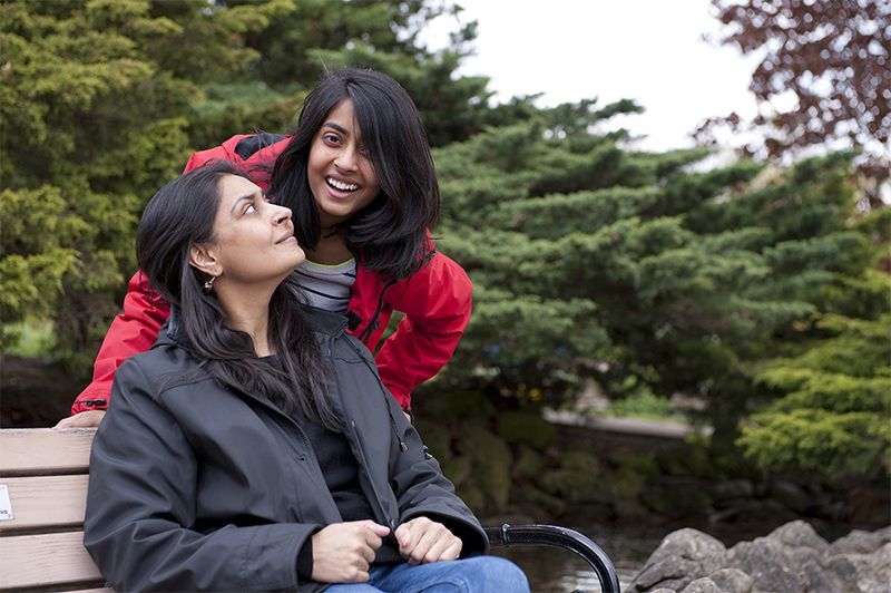 Woman sitting on bench, smiling woman stands behind her in a park setting.