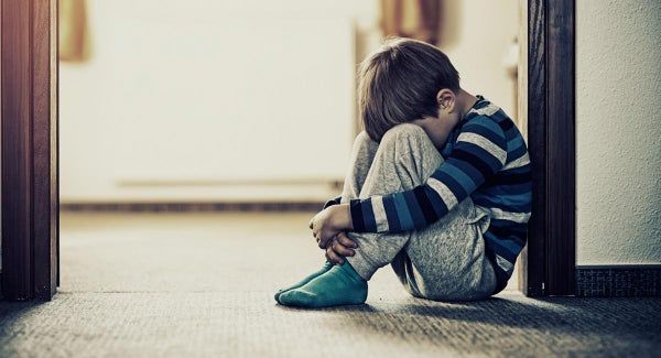 Boy, head in knees, sits in doorway, looking sad. Wearing striped shirt and blue socks. Indoors.