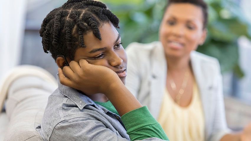 Teen boy looking down, resting head on hand, with woman in background.