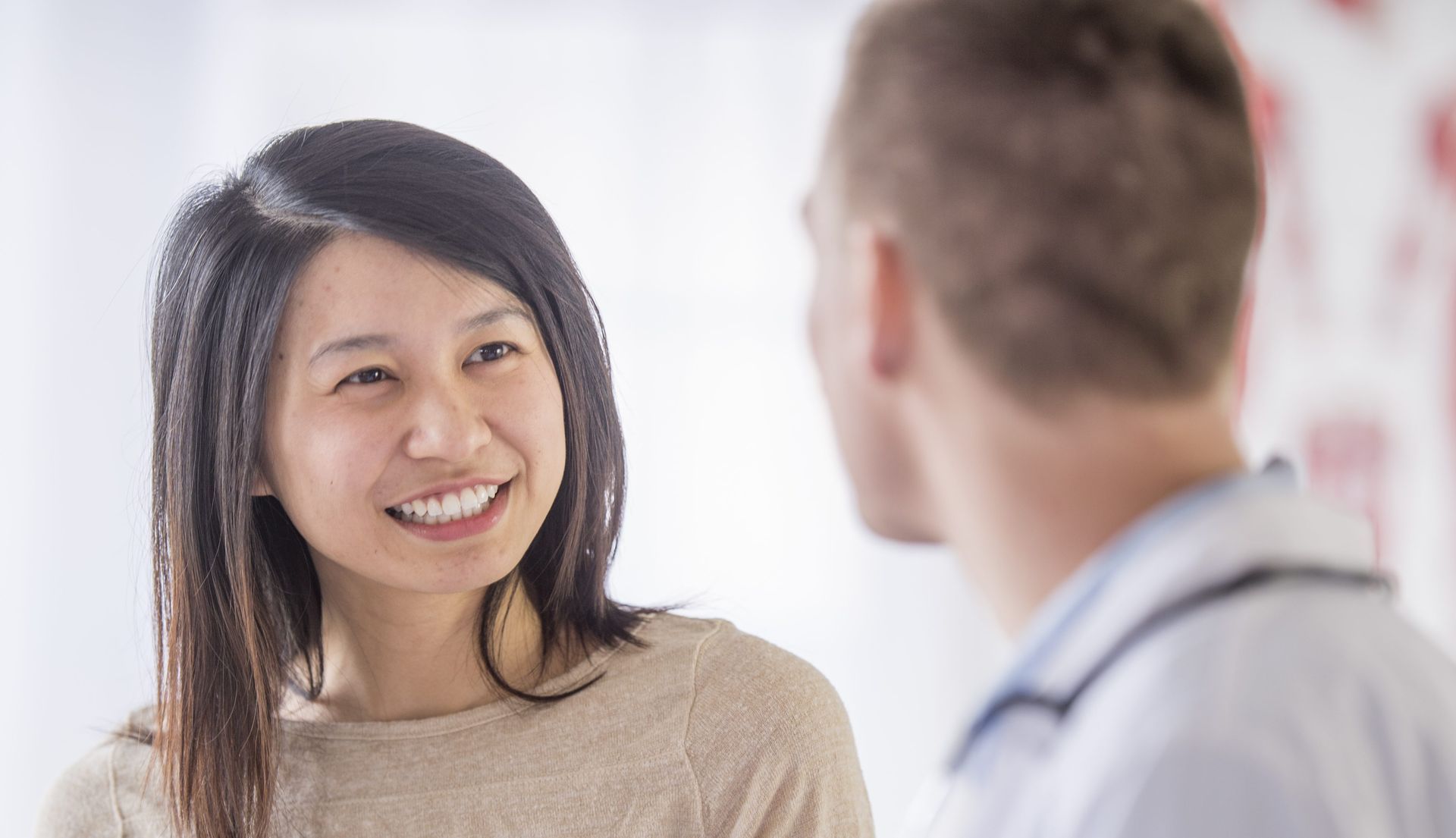 Woman smiling at a doctor in a medical setting; she has black hair and a beige sweater.