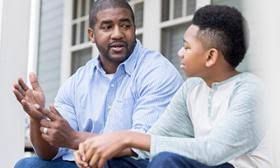 A Black man and teen boy sit on porch, engaged in conversation. The man gestures with his hands.