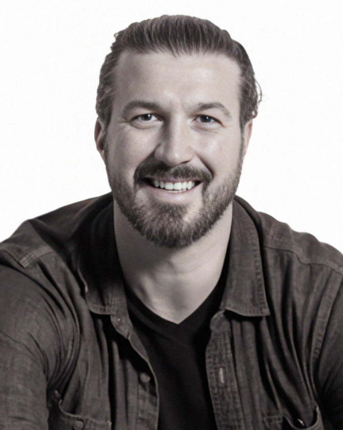 Man with beard smiling, wearing dark shirt and jacket, against white backdrop.
