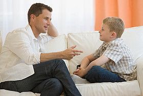 Man in white shirt talking to a young boy on a white couch.