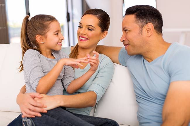 Family of three, girl gesturing, smiling at parents on sofa, indoors.