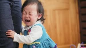 Crying toddler in blue dress, clinging to an adult's leg indoors.