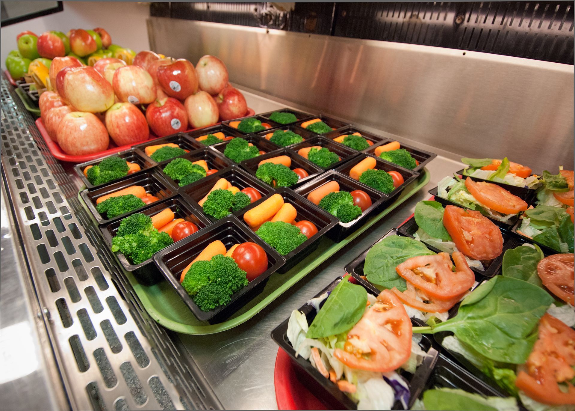 School cafeteria food display: apples, pre-packaged vegetables (broccoli, carrots, tomatoes), and salads.