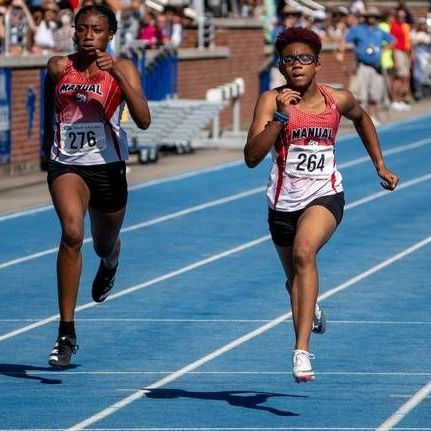 Two female sprinters in red and black uniforms racing on a blue track.