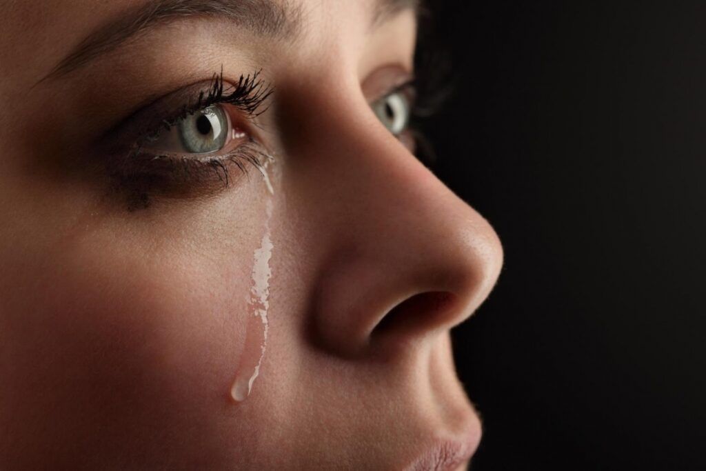 Woman with blue eyes, tear rolling down her cheek, against a black background.