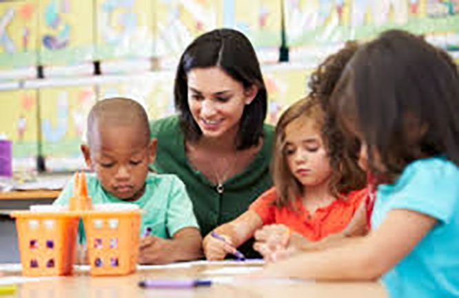 Teacher smiles, helping children draw at a table in a bright classroom.