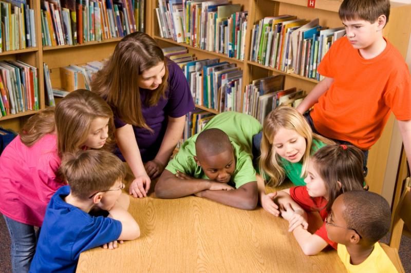 Children and adult in a library looking at something on a table; books on shelves in the background.