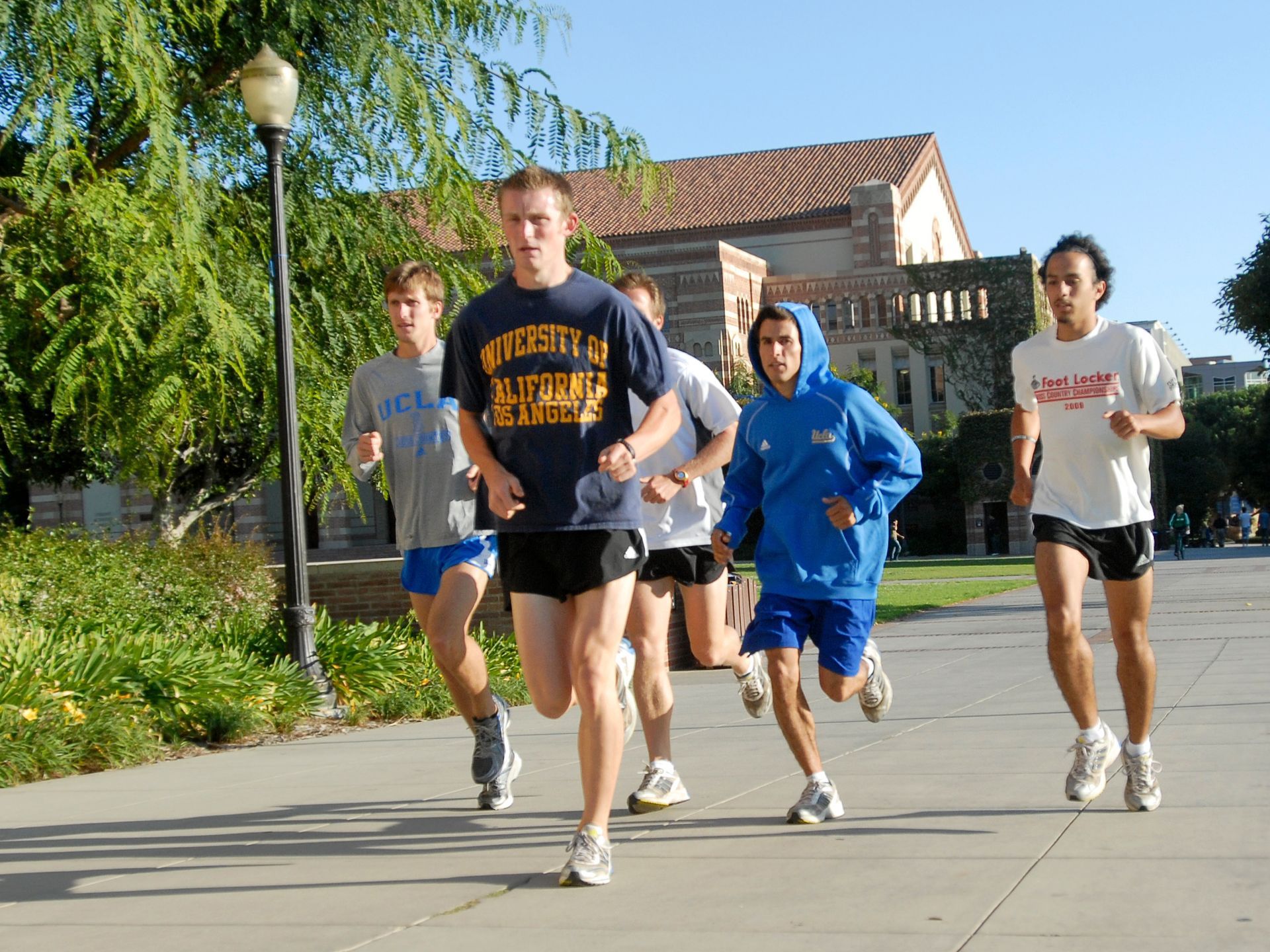 Five young men running on a paved path in front of a building; sunny day.
