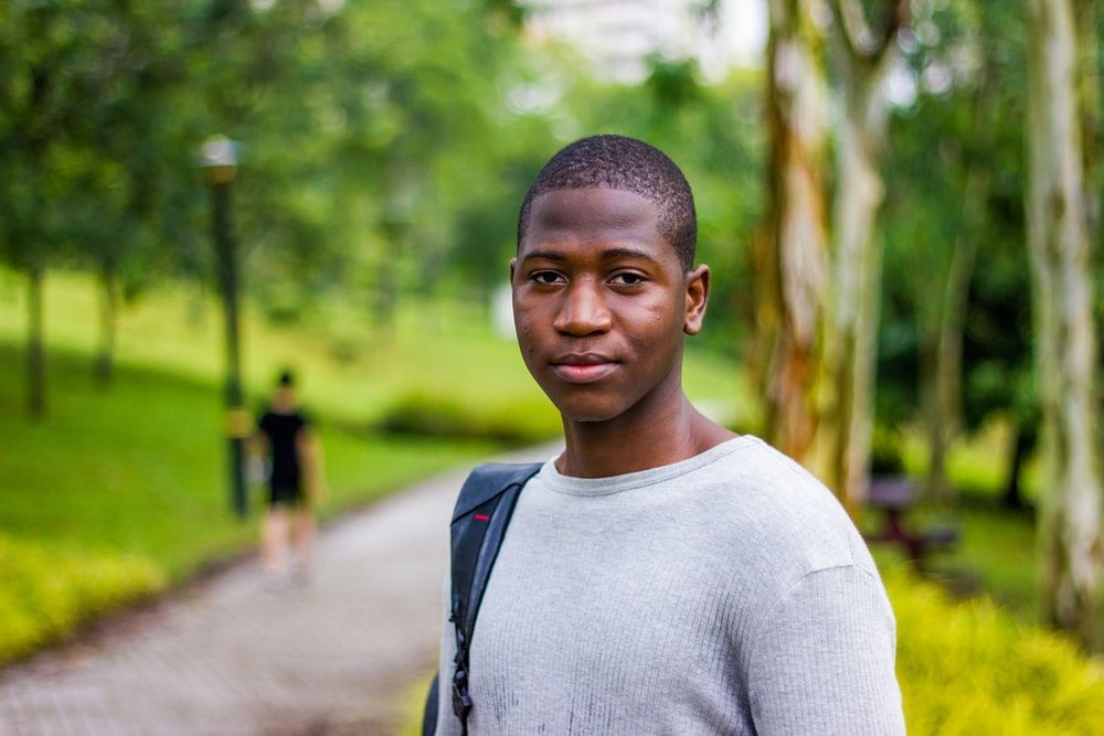 Young Black man with short hair and backpack smiles in a park on a sunny day.