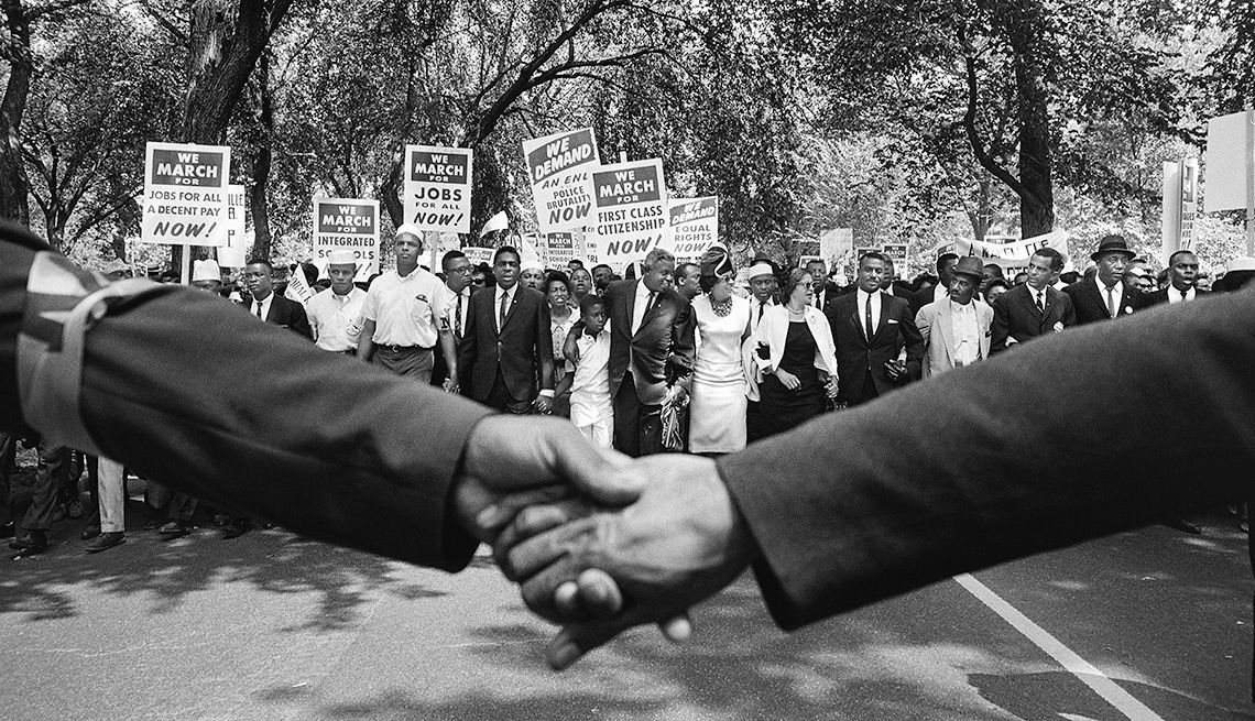 Hands clasp, people march with signs in protest, likely a Civil Rights demonstration.