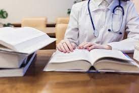 Person in a white lab coat with a stethoscope studies from open books on a desk.