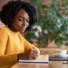Woman in yellow sweater writing in a notebook, with a coffee cup and laptop nearby.