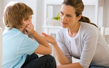 Woman in gray top talking to a boy in a blue shirt; boy covers ears with hands, sitting in a sunny room.