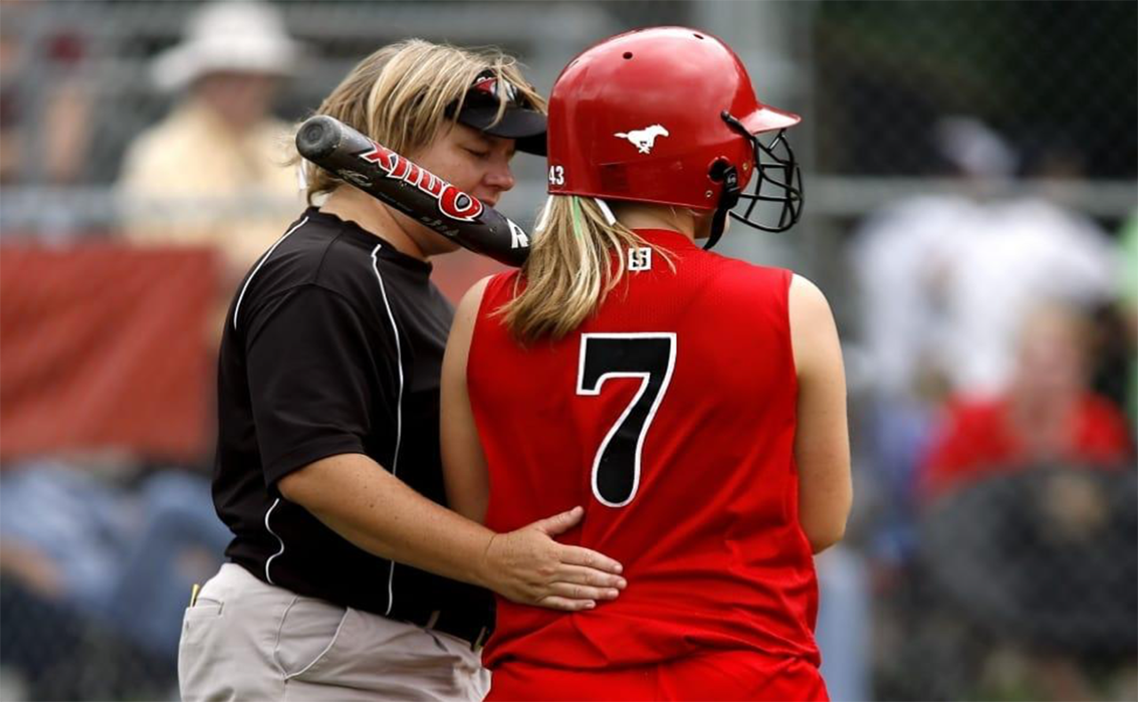 Softball coach with bat and player in red uniform; they are talking on the field.