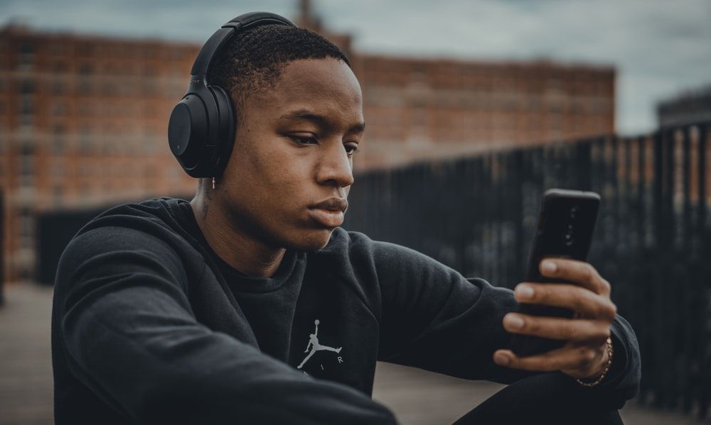 Black man with headphones on, looking at his phone outside.