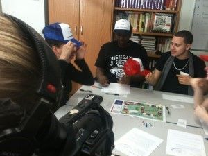 Three people at a table playing a game. One in a black shirt is holding boxing gloves. Bookshelf in background.