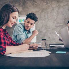 Two people at a table reviewing documents, another person is working on a laptop in the background.
