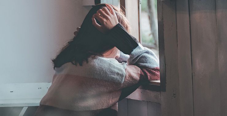 Woman with hands on head, looking out a window. Overcast, neutral tones.